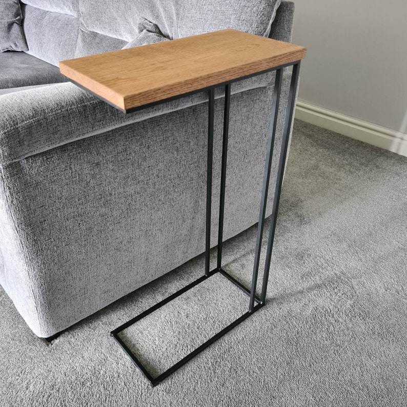 May include: A modern side table with a rectangular, light brown wooden top and a black metal frame. The table is designed to slide alongside a sofa. The table's dimensions are approximately 24 inches long, 12 inches wide, and 24 inches high.