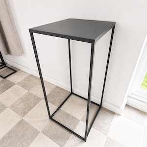 May include: A black metal display stand with a square top and a rectangular base. The stand has a minimalist design, with a sleek, modern aesthetic. The stand is suitable for displaying plants, sculptures, or other decorative items. The height is approximately 75 cm.