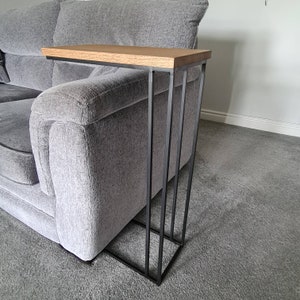 May include: A modern side table with a rectangular oak top and a black metal frame. The table is designed to slide alongside a sofa. The frame has a minimalist design with vertical supports and a square base.