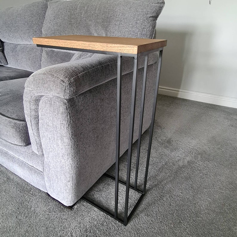 May include: A modern side table with a rectangular oak top and a black metal frame. The table is designed to slide alongside a sofa. The frame has a minimalist design with vertical supports and a square base.