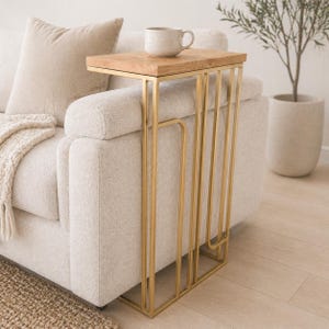 May include: A sofa side table with a light wood top and a gold metal frame. The table is designed to sit alongside a sofa. A beige mug rests on the table's surface. The table is a stylish accent piece for a living room.