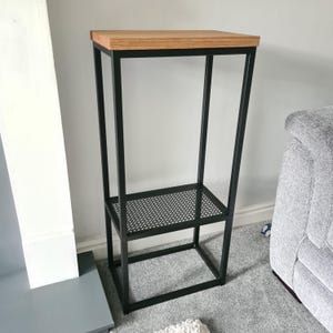 May include: A tall rectangular plant stand featuring a black metal frame with a thin, light brown wooden top surface and a lower shelf made of black metal mesh. The structure is open on all four sides, providing a clear view of the frame.