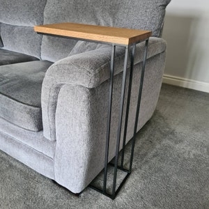 May include: A modern side table with a light brown wooden top and a black metal frame. The table is designed to slide over the arm of a gray sofa. The table's dimensions are approximately 20 inches long, 10 inches wide, and 24 inches high.