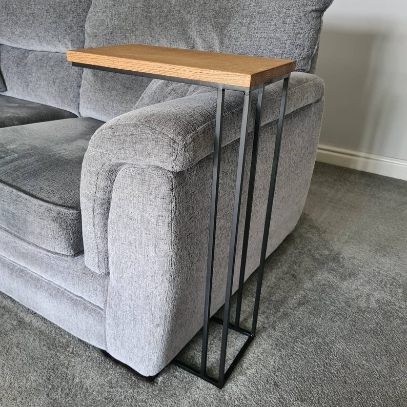 May include: A modern side table with a light brown wooden top and a black metal frame. The table is designed to slide over the arm of a gray sofa. The table's dimensions are approximately 20 inches long, 10 inches wide, and 24 inches high.
