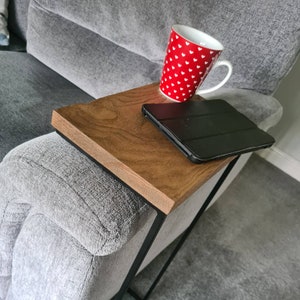May include: A wooden sofa arm table with a dark brown finish and black metal frame. A red mug with white hearts and a black tablet rest on the table. The table is placed on the arm of a grey sofa.