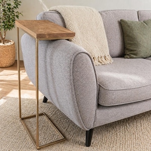 May include: A side table with a rectangular, light brown wooden top and a gold-coloured metal frame. The table is positioned next to a grey sofa with a cream-coloured throw blanket and a green cushion.