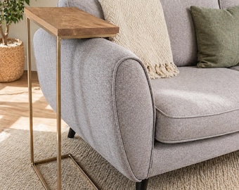 Può includere: Un tavolino con un piano rettangolare in legno marrone chiaro e una struttura in metallo dorato. Il tavolino è posizionato accanto a un divano grigio con una coperta color crema e un cuscino verde.