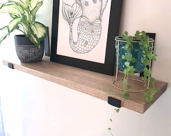 Può includere: Uno scaffale in legno con staffe in metallo nero, che tiene una stampa incorniciata di una sirena con testa d'orso e due piante in vaso. Una pianta è una pianta verde a foglia in un vaso bianco e nero, e l'altra è una pianta verde a foglia in un vaso di vetro blu-verde.