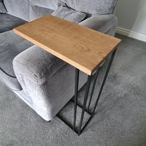 May include: A modern side table with a rectangular, light brown wooden top and a black metal frame. The table is designed to slide over the arm of a sofa. The frame has a geometric, angled design.