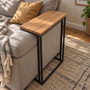 May include: A C-shaped side table featuring a rectangular light brown wood grain top and a slim black metal frame. The table is designed to slide over the arm of a sofa, providing a flat surface for items while maintaining a narrow profile.