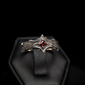 Puede incluir: Un anillo de plata con un diseño intrincado que presenta una piedra preciosa roja en el centro.