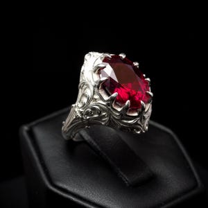 Puede incluir: Un anillo plateado con una gema roja oscura, ovalada y facetada. El anillo tiene un diseño ornamental y arremolinado alrededor de la gema y en la banda. El anillo se exhibe sobre una superficie negra sobre un fondo negro.