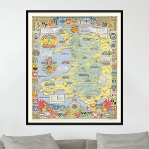 May include: A framed vintage map of Wales with a colorful design featuring coats of arms and other details. The map is surrounded by a black frame and is hanging on a white wall above a gray couch.