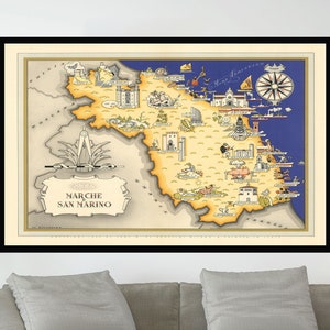May include: A vintage map of Marche and San Marino, Italy, with a compass rose and illustrations of buildings and landmarks. The map is framed in black and displayed on a wall above a gray couch.