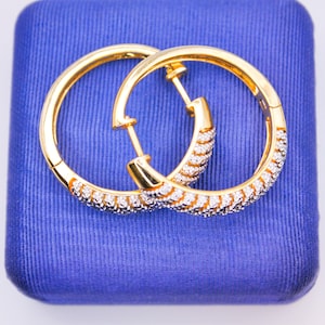 May include: A pair of gold hoop earrings with a row of small clear stones around the edge. The earrings are on a blue velvet background.