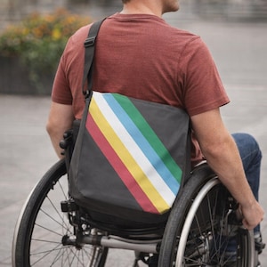 May include: A dark gray messenger bag with a diagonal striped pattern in red, yellow, white, light blue, and green. The bag features an adjustable black shoulder strap and is worn across the back, resting against a maroon t-shirt and a wheelchair.
