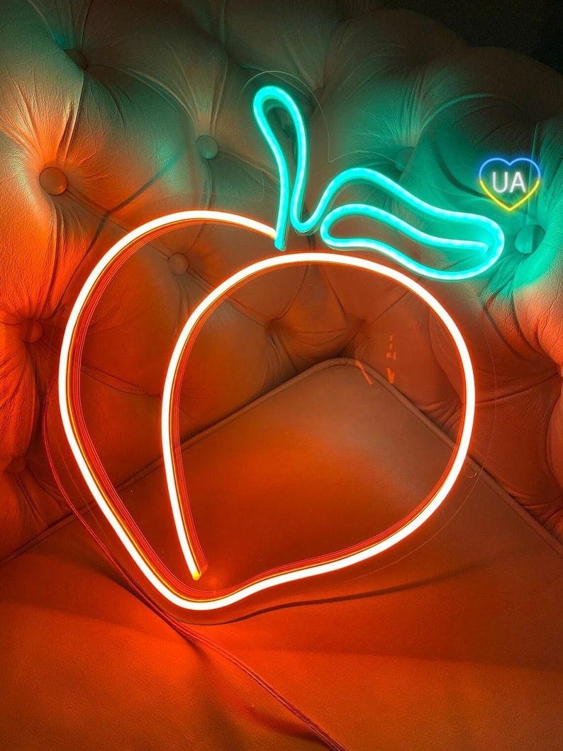 Puede incluir: Un letrero de melocot&oacute;n de ne&oacute;n con un tallo y una hoja verdes. El letrero est&aacute; iluminado y brilla en naranja. El letrero est&aacute; en un sof&aacute; marr&oacute;n con un coraz&oacute;n con las letras "UA" dentro.