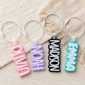 May include: Four silicone luggage tags with white raised lettering on colored backgrounds. The tags read Olivia in pink, Noah in light purple, Madison in black, and Emma in teal. Each tag is attached to a flexible silver-toned metal cable loop.
