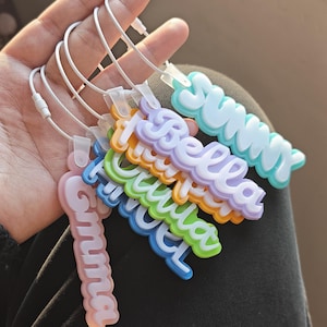 Op de afbeelding: Een verzameling kleurrijke naamlabels met witte draadlussen. De labels tonen namen als "Sunny", "Bella" en "Emma" in een speels, bubble-lettertype. Elk label heeft een andere kleur, waaronder roze, blauw, groen, oranje en paars.