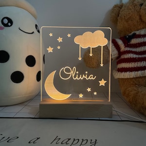 May include: A white acrylic night light with a gold crescent moon, cloud, and stars. The name "Olivia" is written in gold script on the light. The light is sitting on a wooden base.
