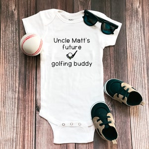 May include: White baby onesie with black text that reads "Uncle Matt's future golfing buddy" with a golf club and ball graphic. A baseball and a pair of baby shoes are on a wood background.