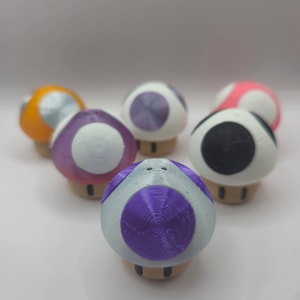 May include: A collection of six 3D printed mushroom-shaped figurines. The mushrooms are white with different coloured tops, including purple, orange, black, and pink.