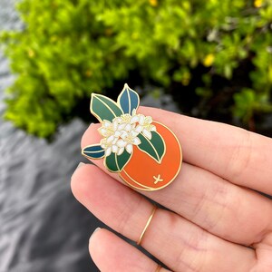 May include: Gold enamel pin featuring a stylized orange with white blossoms and green leaves. The orange is half-sliced and shows a cross inside.