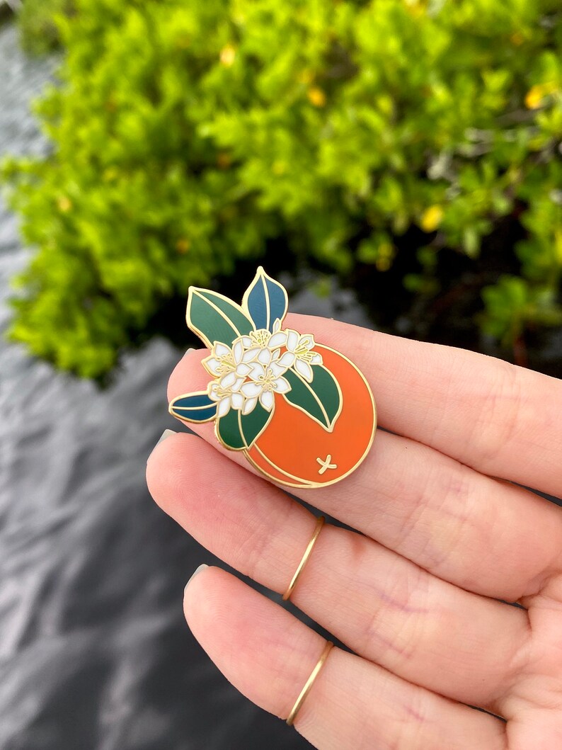 May include: Gold enamel pin featuring a stylized orange with white blossoms and green leaves. The orange is half-sliced and shows a cross inside.