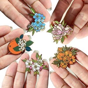 May include: Five enamel pins featuring different floral designs. The pins are gold-toned and feature a variety of flowers, including forget-me-nots, peonies, orange blossoms, apple blossoms, and California poppies.