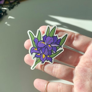 May include: A purple and green iris flower sticker with a white background.