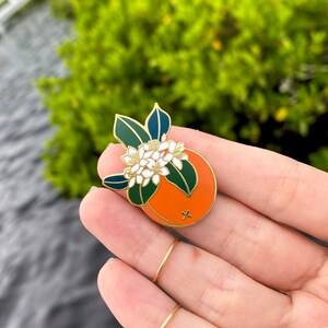 May include: Gold enamel pin featuring a stylized orange with white blossoms and green leaves.