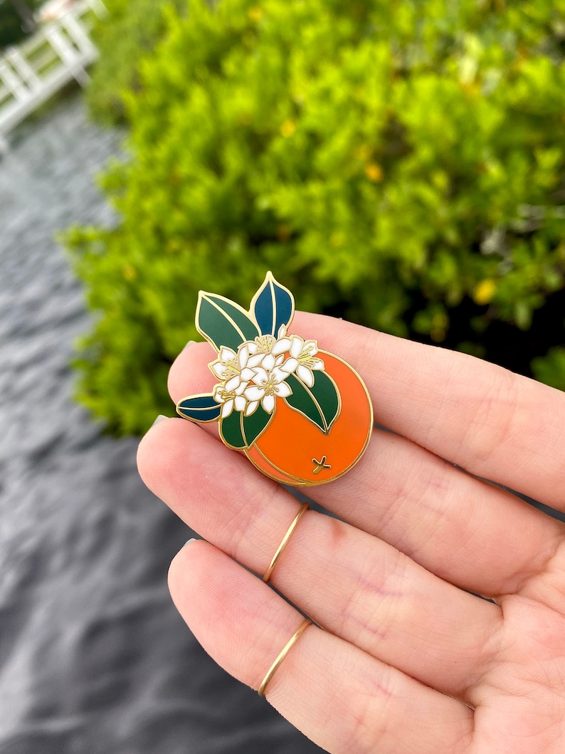 May include: Gold enamel pin featuring a stylized orange with white blossoms and green leaves.