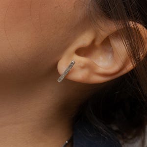Puede incluir: Un pendiente de barra plateada se lleva en la oreja. El pendiente tiene forma rectangular pequeña con una superficie texturizada. La persona lleva una blazer azul oscuro y un collar de cadena plateada.