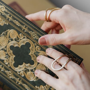May include: Two gold-colored rings are displayed on fingers, one with a double-band design and the other with a teardrop shape. The rings are set against a backdrop of a dark green book with gold decorative accents.