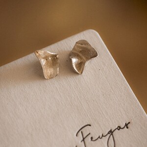 May include: A pair of silver earrings with an organic, asymmetrical shape. The earrings are displayed on a white card with the text "Lucia Feugoas Joyeria de Autor".