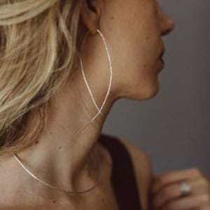 Puede incluir: Pendientes de aro de plata con un diseño delgado y curvo. Los pendientes son grandes y cuelgan de los lóbulos de las orejas.