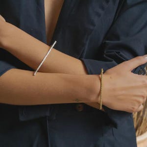 Pode incluir: Uma pessoa vestindo um terno azul escuro usa duas pulseiras. Uma é prateada, a outra dourada. Ambas são finas e lineares. As pulseiras são o ponto focal, adicionando um toque de elegância. A imagem é bem iluminada, destacando as joias.