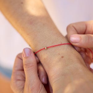 Pode incluir: Uma pulseira de fio vermelho com uma pequena conta dourada. A pulseira está sendo amarrada em volta de um pulso. A imagem é um close-up, destacando o design simples da pulseira e a textura da pele.