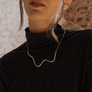 Puede incluir: Un collar plateado con un diseño ondulado y orgánico. El collar se lleva sobre un cuello alto negro. El fondo presenta una pared de ladrillos y una pared blanca.