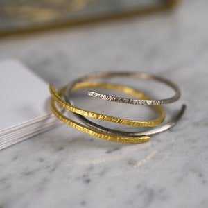 Pode incluir: Três pulseiras finas de ouro e prata com acabamento texturizado. As pulseiras estão empilhadas umas sobre as outras.