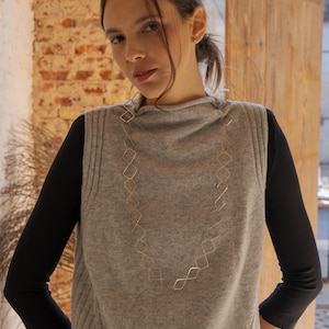 Puede incluir: Una mujer con un suéter sin mangas gris sobre una camisa negra de manga larga. Un collar geométrico dorado cuelga por delante. El fondo incluye una pared de ladrillo y un marco de puerta de madera.