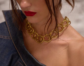 Puede incluir: Un collar de cadena dorado con eslabones circulares grandes e interconectados. El collar se usa contra una chaqueta de mezclilla y una persona con lápiz labial rojo.