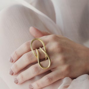 Peut inclure: Une bague dorée au design abstrait. La bague présente une forme continue et bouclée qui s'enroule autour du doigt. La bague est portée sur une main aux ongles manucurés.