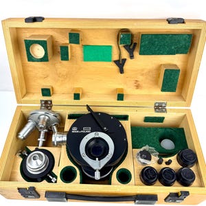 Puede incluir: Maleta de madera abierta que contiene equipo científico. La caja tiene un acabado de madera clara y está forrada con fieltro verde. El equipo incluye un microscopio, lentes y otros componentes. El texto "MADE IN POLAND" es visible.