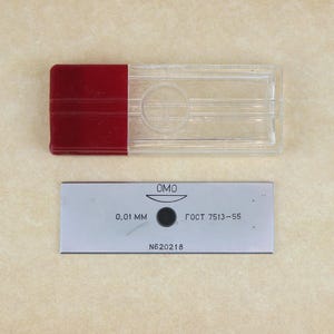 May include: A clear plastic rectangular case with a red lid, and a small rectangular metal plate. The plate has the text "0,01 mm" and other Cyrillic text. The case and plate are on a beige surface.