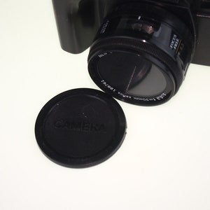 May include: A black camera lens with a 1:3.5 50mm lens and a black lens cap that says "Camera" on it.