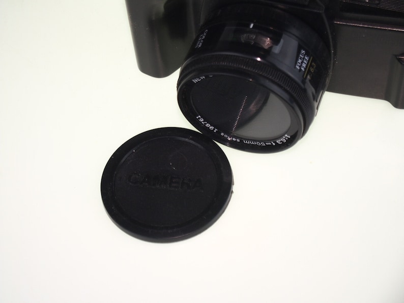 May include: A black camera lens with a 1:3.5 50mm lens and a black lens cap that says "Camera" on it.