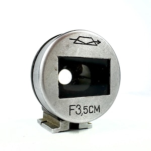 May include: A circular silver-toned camera viewfinder accessory featuring a rectangular glass lens opening. The front face displays a black graphic symbol of a rectangle with an arrow and the printed text F3,5CM. It includes a metal mounting base.