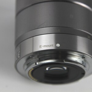 Puede incluir: Un objetivo de c&aacute;mara plateado con una tapa de lente negra y el texto "E-mount" impreso en el lateral.