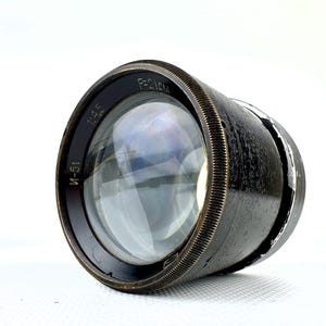 Könnte beinhalten: Ein Vintage-Kameraobjektiv mit schwarzem und bronzefarbenem Finish. Das Objektiv hat ein großes, klares Glaselement und ist mit "1:4.5" und "F=21CM" gekennzeichnet. Das Objektiv hat einen geriffelten, bronzefarbenen Ring am Rand.